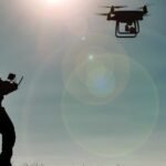 Silhouette of a person operating a flying drone against a bright sky.