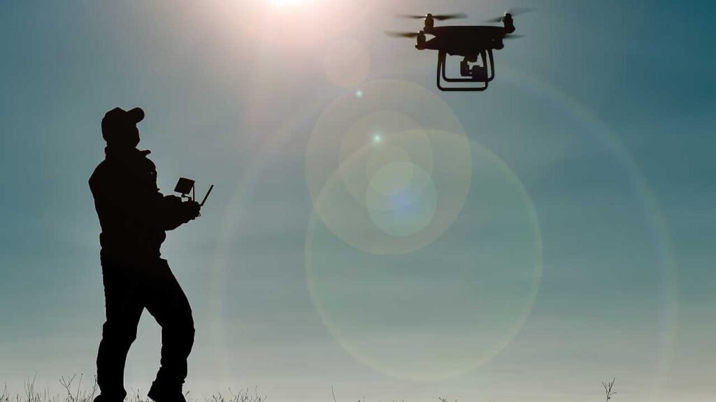 Silhouette of a person operating a flying drone against a bright sky.