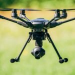 A black drone with six propellers hovers in the air against a blurred green and yellow background.