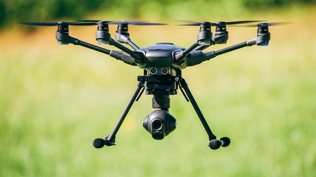 A black drone with six propellers hovers in the air against a blurred green and yellow background.