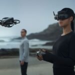 Two people operate a drone on a beach. One wears virtual reality goggles and holds a control device, while the other watches the drone. Waves and cliffs are visible in the background.