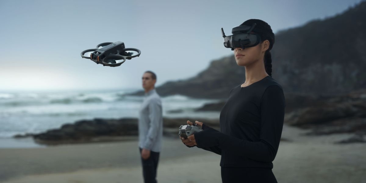 Two people operate a drone on a beach. One wears virtual reality goggles and holds a control device, while the other watches the drone. Waves and cliffs are visible in the background.