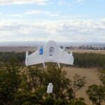 A white drone flying over a rural landscape with trees and fields, carrying a small package underneath.