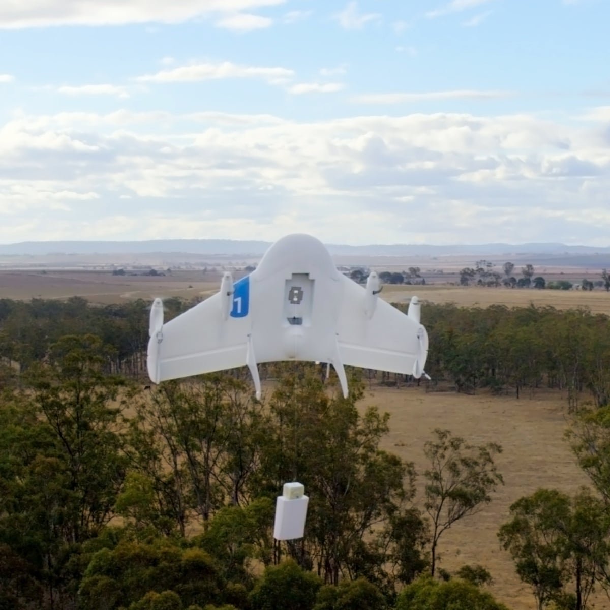 A white drone flying over a rural landscape with trees and fields, carrying a small package underneath.