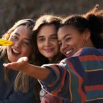 Three people sit together, smiling and posing for a group selfie outdoors.