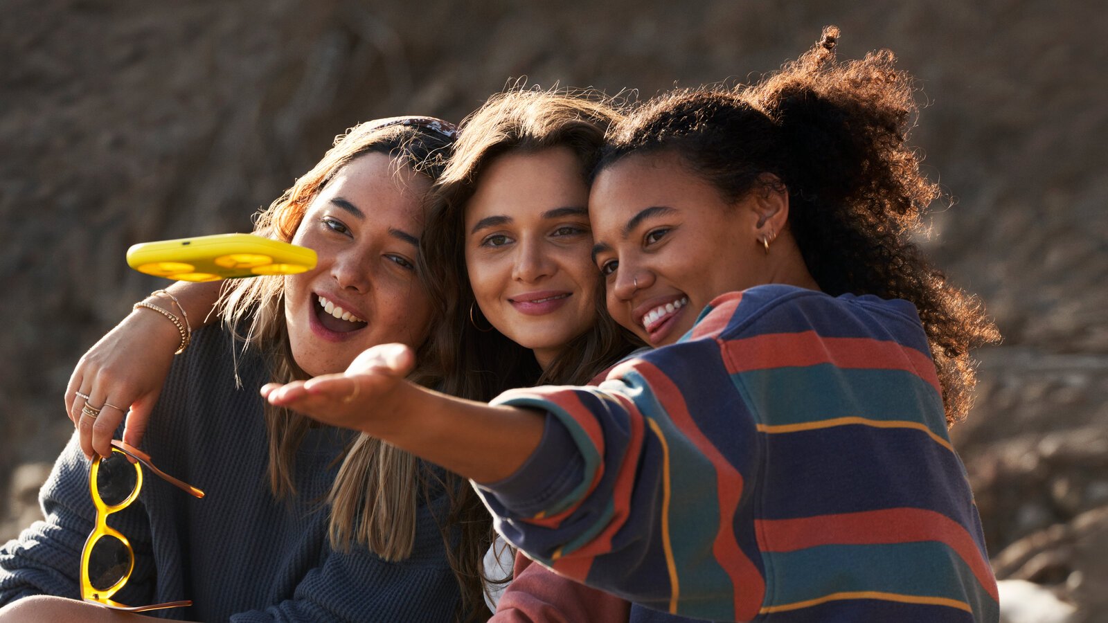 Three people sit together, smiling and posing for a group selfie outdoors.