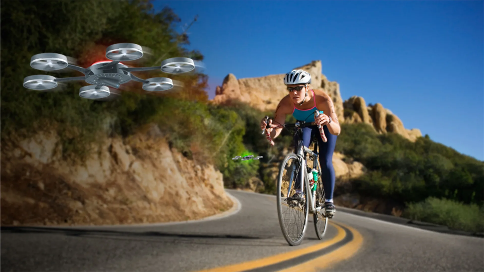 Cyclist in gear rides on a winding road between hills, while a large drone with multiple propellers hovers nearby.