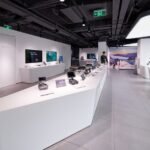 A modern store interior with sleek white tables displaying electronic devices, screens on the walls, and bright overhead lighting. A person is browsing the products.