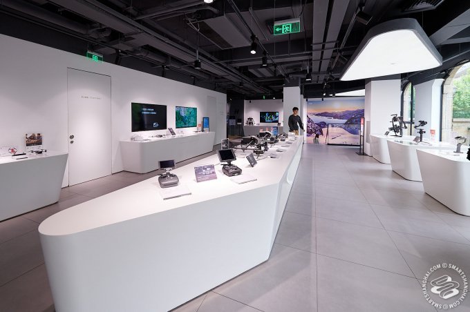 A modern store interior with sleek white tables displaying electronic devices, screens on the walls, and bright overhead lighting. A person is browsing the products.