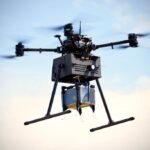 A delivery drone in flight carrying a package beneath it against a clear sky.