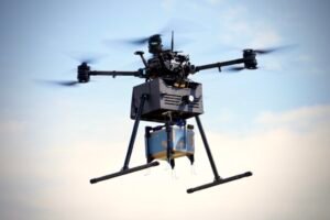 A delivery drone in flight carrying a package beneath it against a clear sky.