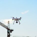 A drone hovers in the sky, with a construction crane visible in the background against a clear blue sky.