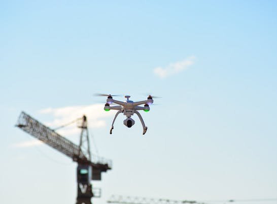 A drone hovers in the sky, with a construction crane visible in the background against a clear blue sky.