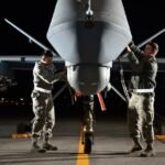 Two technicians work on the nose of a military drone on an airstrip at night.