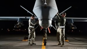 Two technicians work on the nose of a military drone on an airstrip at night.