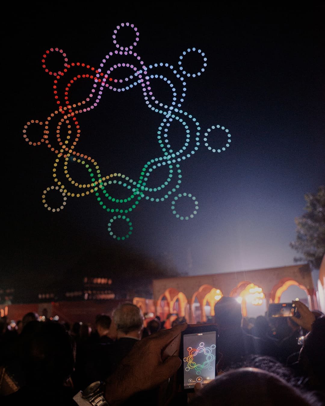 Colorful circular pattern of lights in the night sky above a crowd, with some people taking photos.