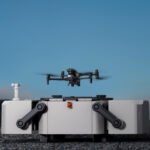 A drone hovers above a docking station on a rocky landscape with mountains in the background under a clear sky.