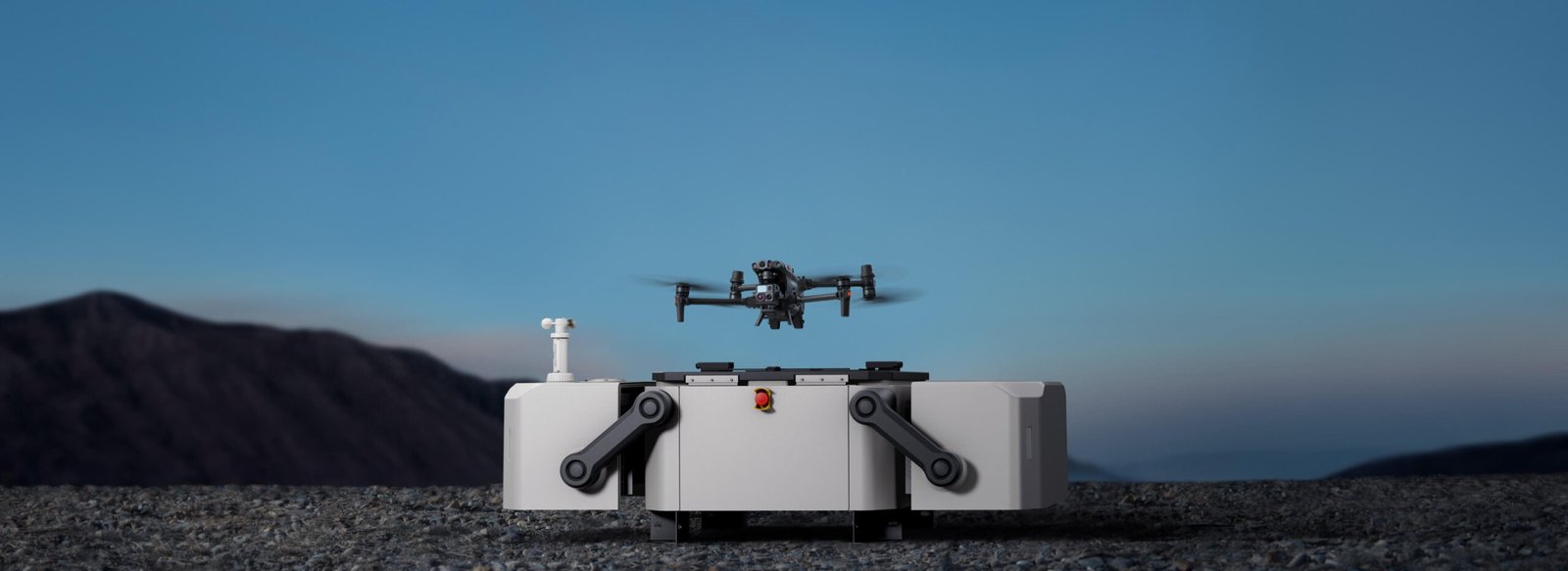 A drone hovers above a docking station on a rocky landscape with mountains in the background under a clear sky.