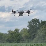 A drone flies over a cornfield with trees in the background under a cloudy sky.