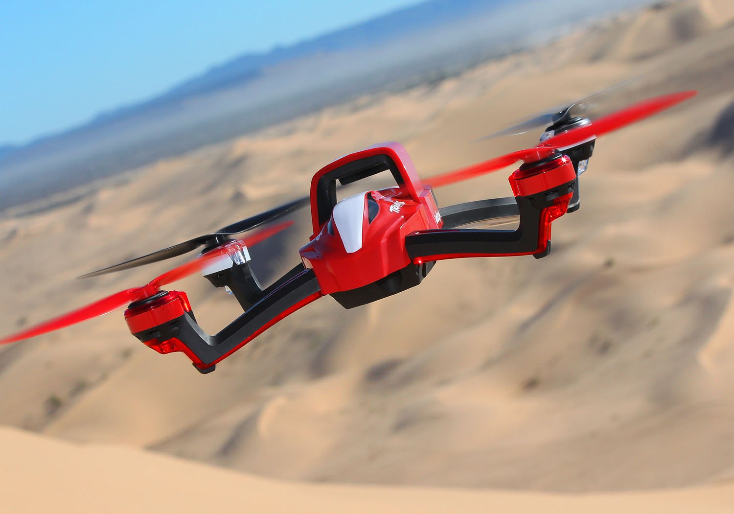 A red and black drone flies over desert sand dunes under a clear blue sky.