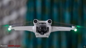 A white drone flying with its camera facing forward, lights on its arms glowing.