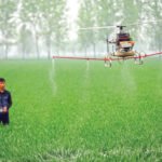 A person operates a drone spraying pesticide over a lush green field with trees in the background.