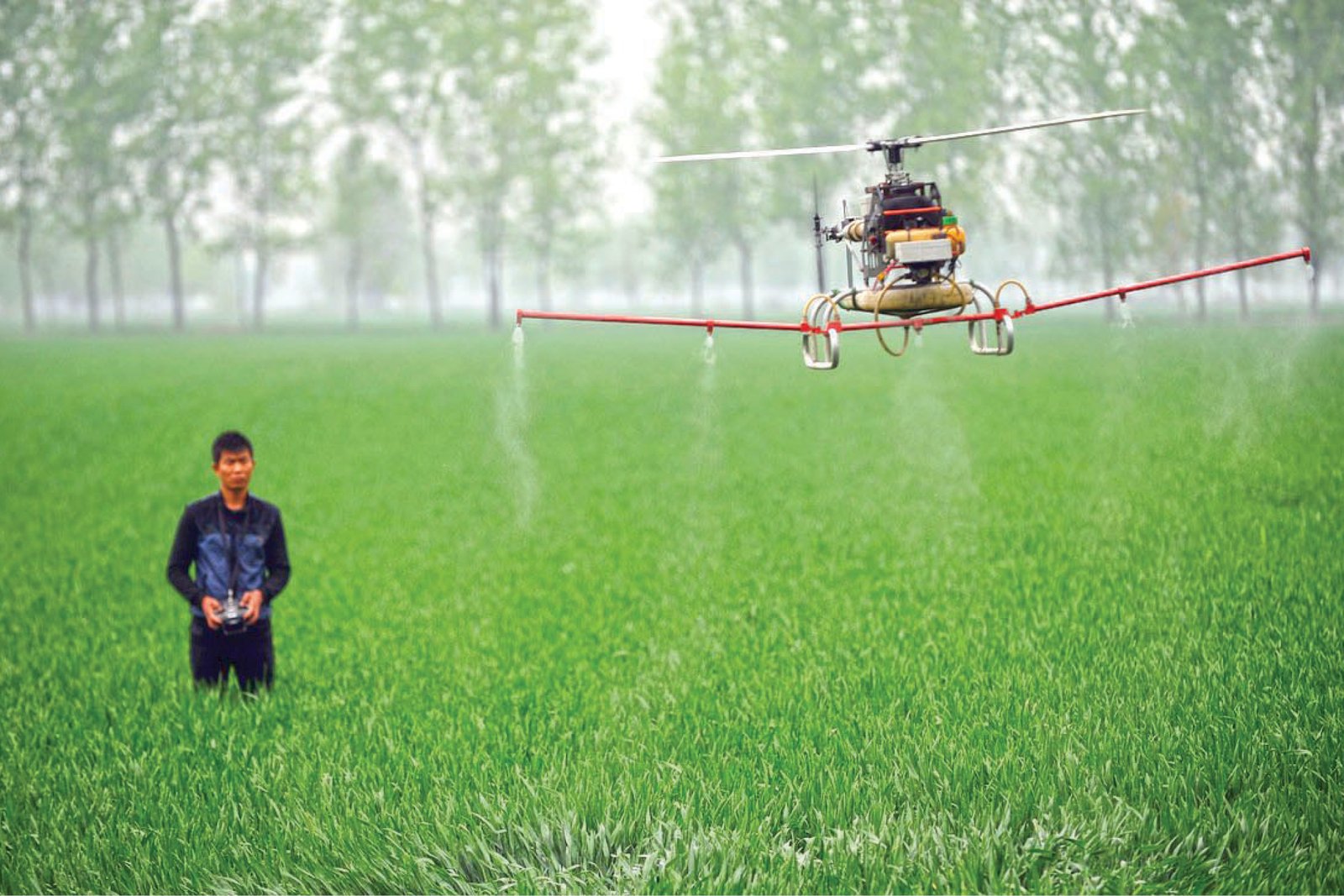 A person operates a drone spraying pesticide over a lush green field with trees in the background.