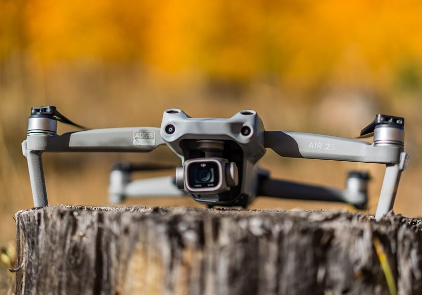 A gray drone with a camera sits on a tree stump against a blurred autumnal background.