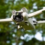 A small, white drone with four rotors hovers in the air against a backdrop of green foliage.