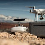 A white drone and its remote control rest on a rocky surface with mountains and a clear blue sky in the background.