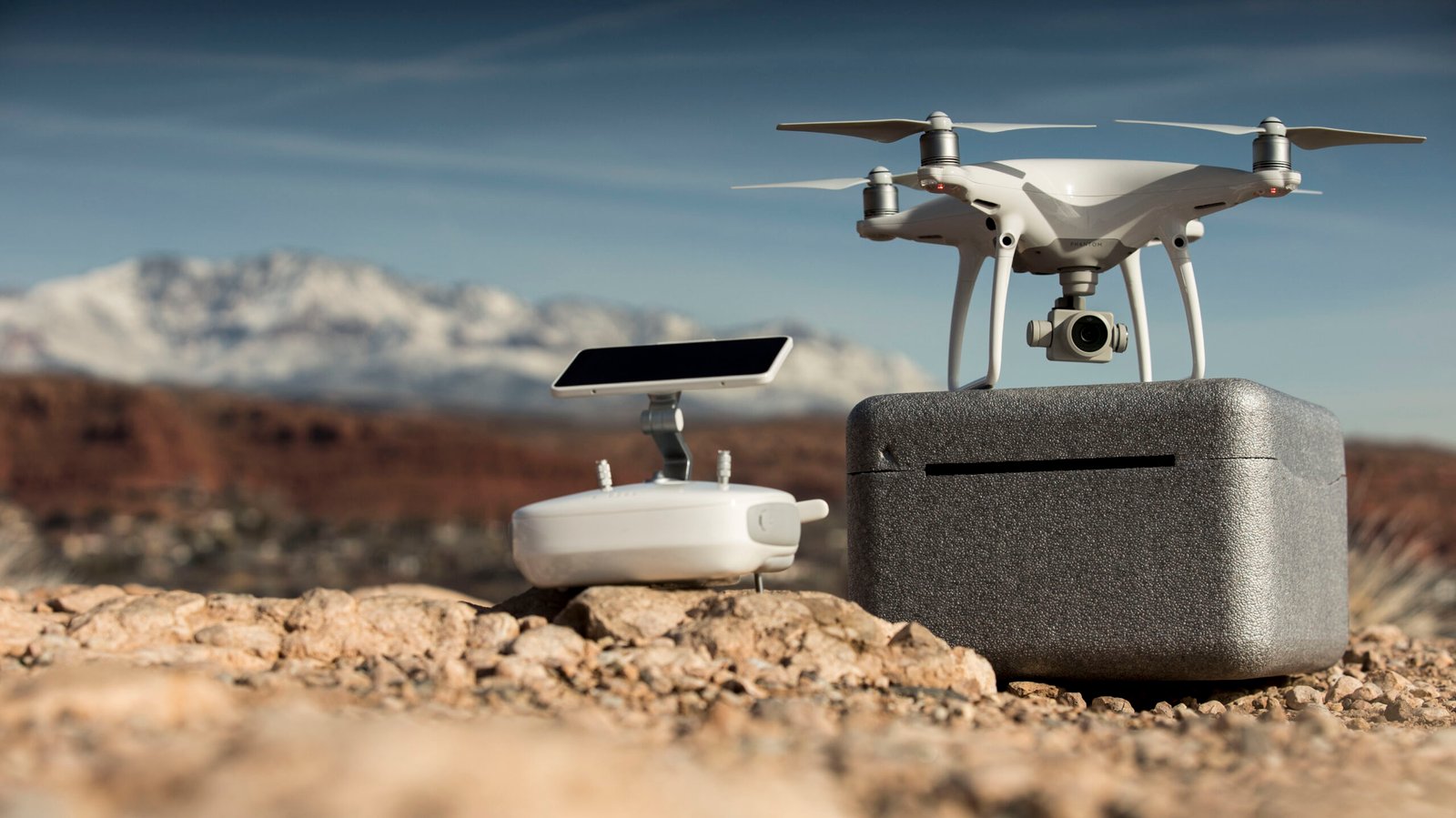A white drone and its remote control rest on a rocky surface with mountains and a clear blue sky in the background.