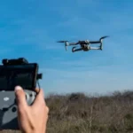 A person operates a drone with a remote control, flying over an open field under a clear blue sky.