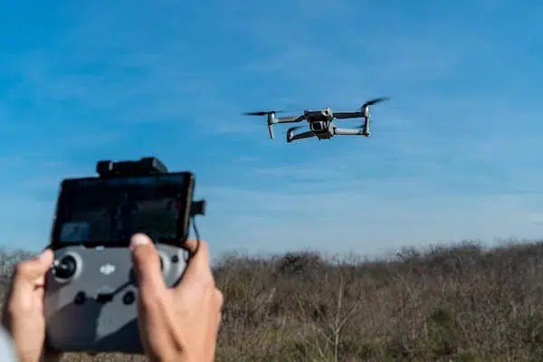 A person operates a drone with a remote control, flying over an open field under a clear blue sky.