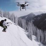 Snowboarder on a snowy mountain slope being recorded by a drone, surrounded by snow-covered trees and mountains.