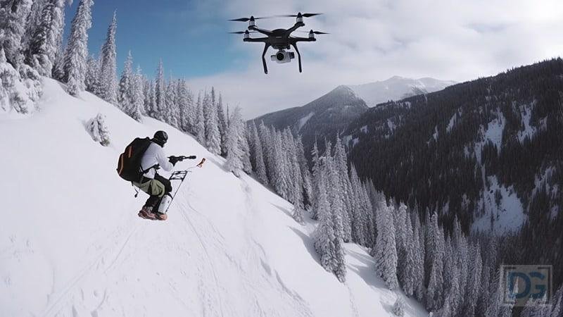 Snowboarder on a snowy mountain slope being recorded by a drone, surrounded by snow-covered trees and mountains.