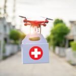 A drone delivering a white box with a red medical cross flies down a suburban street.