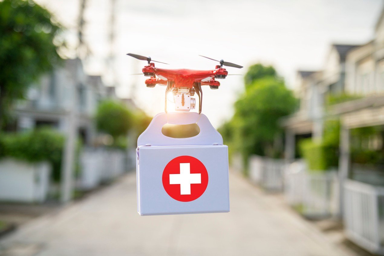 A drone delivering a white box with a red medical cross flies down a suburban street.