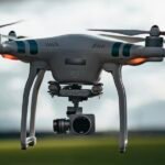 A white drone equipped with a camera is flying outdoors against a blurred background of sky and greenery.