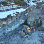 Aerial view of construction site with machinery on gravel, surrounded by snow and rocky terrain.