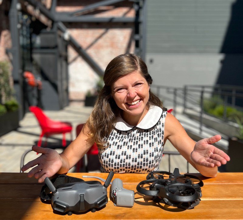 A woman sits at a table outdoors with drone equipment in front of her, smiling at the camera.