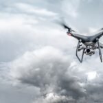 A drone with a camera flies against a cloudy sky backdrop.