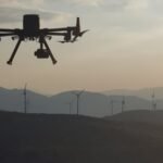 A drone hovers in the sky at sunset, with wind turbines visible in the background against silhouetted hills.