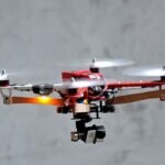 A red and white drone with four propellers hovers in the air against a gray background.