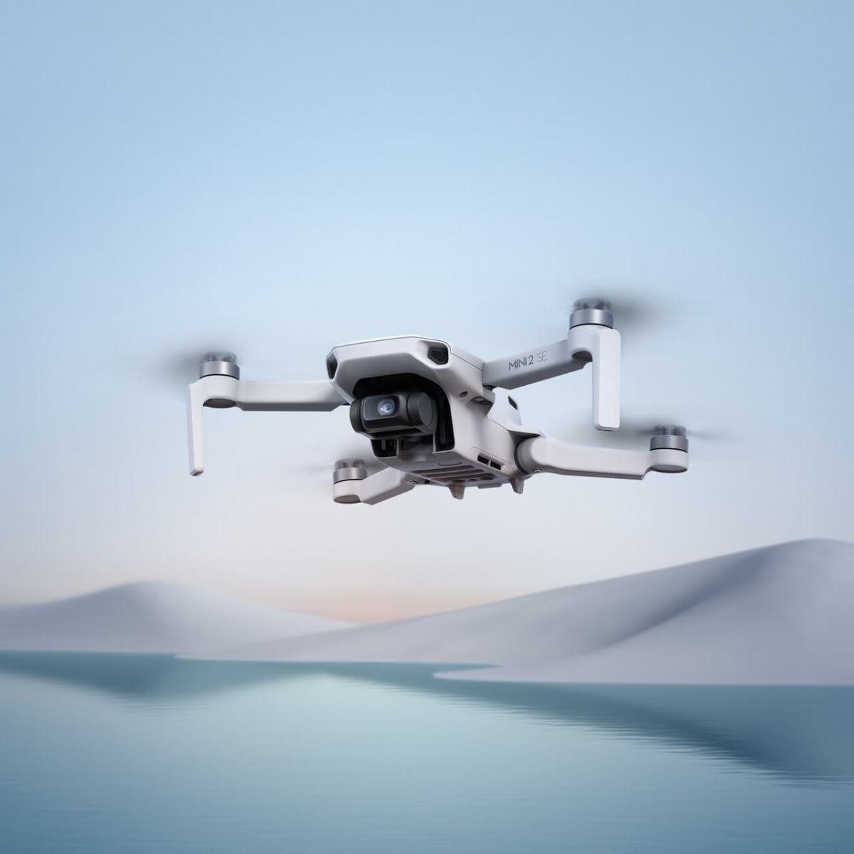 A white drone hovers above a serene landscape with blue water and soft, blurred hills in the background.