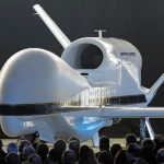 A large drone aircraft displayed indoors with a crowd of people observing it.