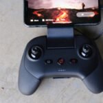 Black game controller connected to a smartphone displaying a game scene on a gray surface.