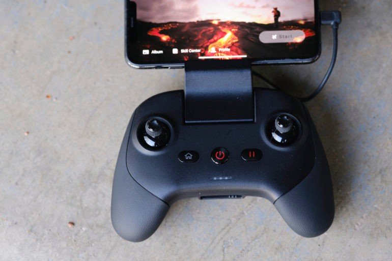 Black game controller connected to a smartphone displaying a game scene on a gray surface.