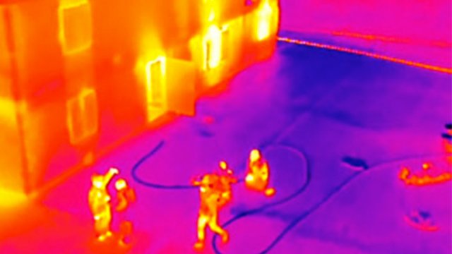 Thermal image of several figures outside a building, possibly firefighters, with hoses and heat variations shown in different colors.