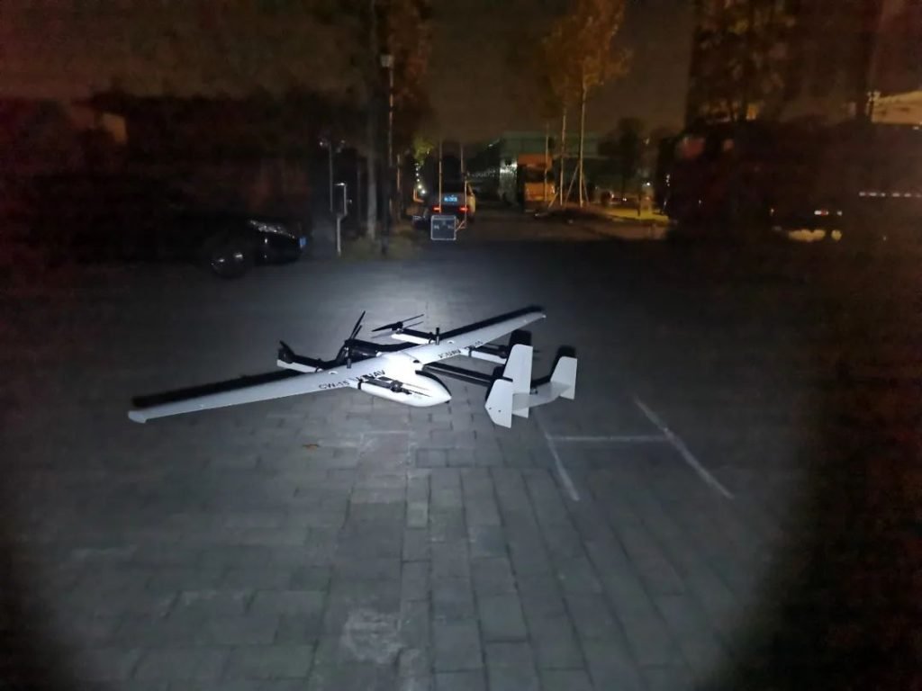 A large drone is illuminated on the ground in a dark outdoor area with faint buildings and parked vehicles in the background.