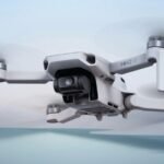 A close-up view of a DJI Mini 2 SE drone in flight against a blurred sky and water background.